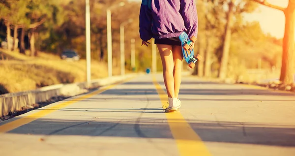 Стильная молодая девушка в фиолетовой куртке идет по дороге держа скейтборд в руке — стоковое фото