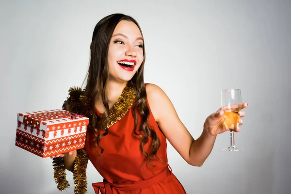 Glad ung flicka i röd klänning firar nyår och jul, håller ett glas champagne och en gåva — Stockfoto