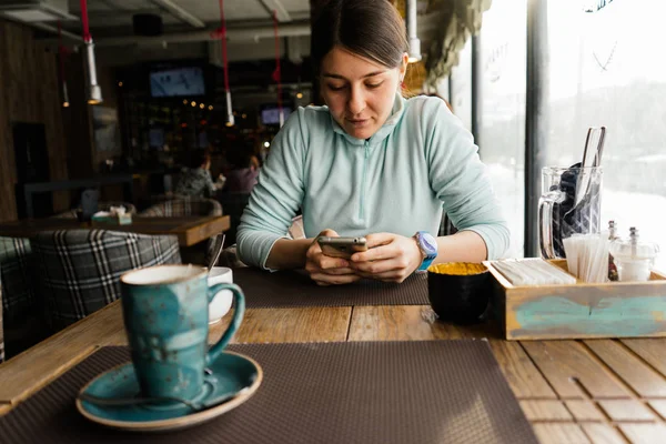 Linda chica joven en un suéter azul se sienta en un café y espera su comida, mira en un teléfono inteligente —  Fotos de Stock