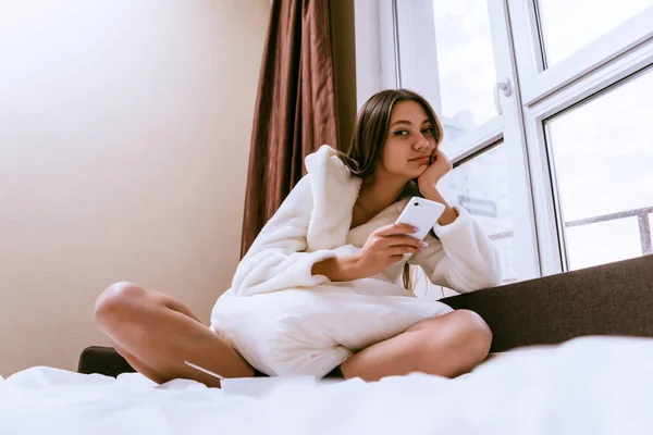 Задумчивая женщина сидит на кровати и держит телефон в руках — стоковое фото
