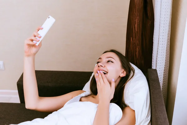 Wanita bahagia berbaring di bawah selimut tersenyum dan melihat telepon — Stok Foto