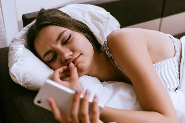 Wanita mengantuk di tempat tidur di bawah selimut memegang telepon di tangan — Stok Foto