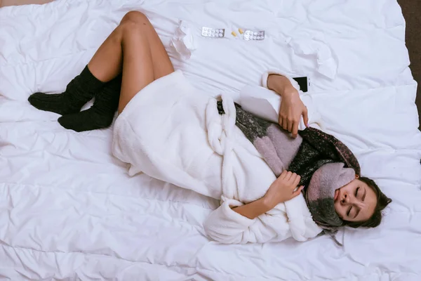Sick woman in a warm scarf is lying on the bed — Stock Photo, Image