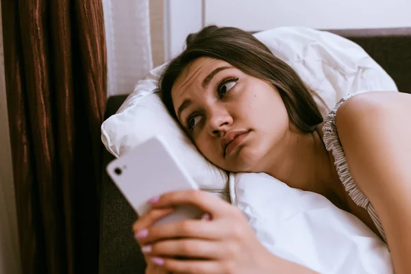 Wanita sedih berbaring di tempat tidur di bawah selimut dan memegang telepon di tangannya — Stok Foto