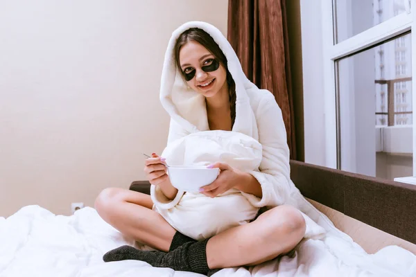 Uma mulher feliz em um casaco branco colocar manchas sob os olhos e senta-se na cama — Fotografia de Stock