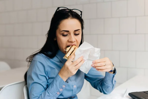 Wanita lapar duduk di kantor di meja dan makan sandwich — Stok Foto