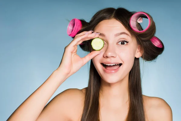 Mulher feliz com encrespadores na cabeça segura um pepino perto de seu olho — Fotografia de Stock