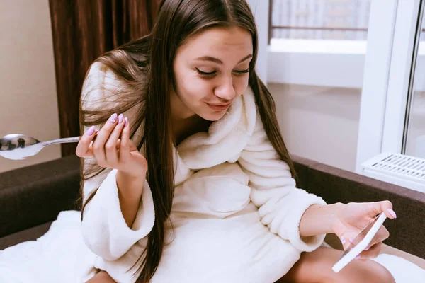 Mulher está sentada na cama tomando café da manhã e olhando para o telefone — Fotografia de Stock