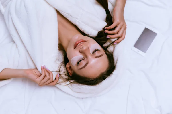 Eine Frau im Mantel liegt im Bett und legt sich Pflaster unter die Augen — Stockfoto