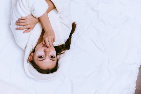 Uma mulher em um manto está deitada na cama e colocar manchas sob os olhos — Fotografia de Stock