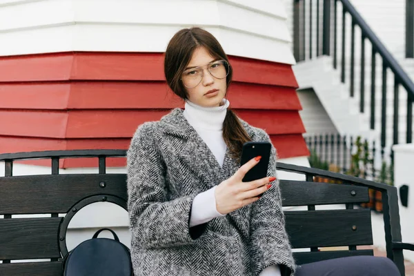 Belle jeune fille urbaine brune avec des lunettes et un manteau gris assis sur un banc dans les rues de la ville, faire des selfies — Photo