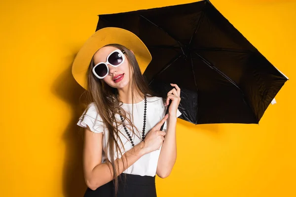 Modelo de chica hermosa con estilo en sombrero de moda y gafas de sol posando sobre fondo naranja sosteniendo un paraguas — Foto de Stock