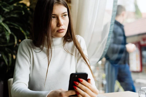 Dark-haired pretty girl sitting in a cafe, holding a smartphone, looking thoughtfully out the window — Stock Photo, Image