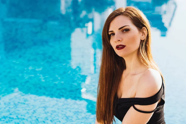 Sexy de pelo largo hermosa chica en un vestido negro se sienta junto a la piscina azul, disfruta de unas vacaciones de lujo — Foto de Stock