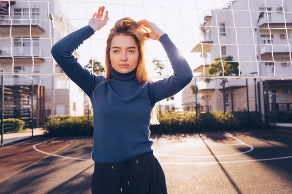 Stijlvolle mooi blond meisje in een blauwe trui poseren op een voetbalveld buiten in de zon — Stockfoto