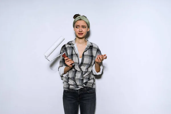 Ontevreden meisje in een shirt houdt een roller voor schilderen muren, moe van het doen van reparaties — Stockfoto