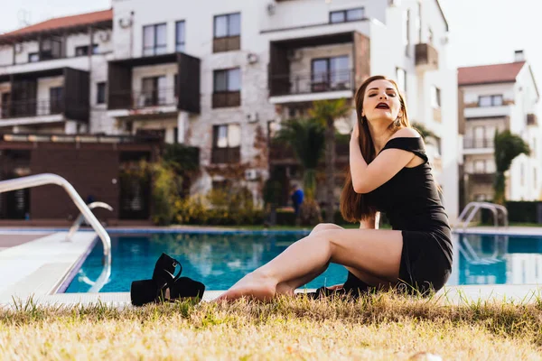 Sexy dai capelli lunghi bella ragazza in un abito nero si siede vicino alla piscina blu, gode di riposo di lusso e una vita ricca — Foto Stock