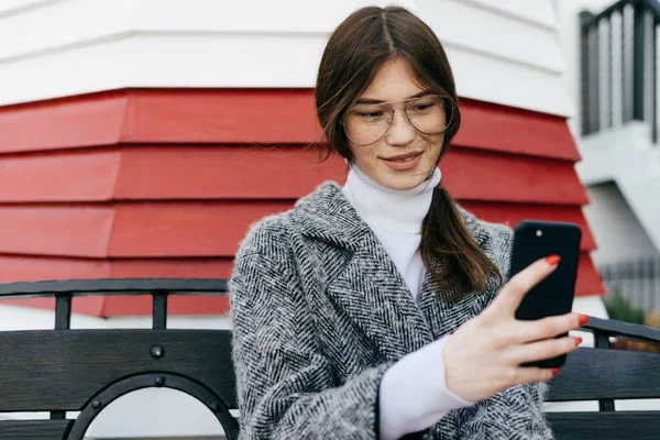 A nice dark-haired girl with glasses and a gray coat is sitting on a bench in the open air, making selfies — Stock Photo, Image