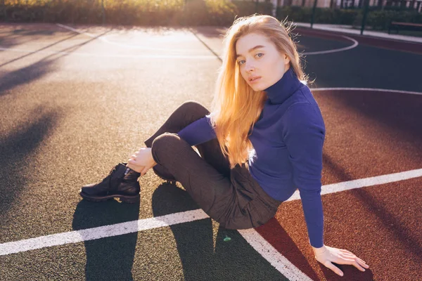 Mooie stijlvolle jonge blonde model in blauwe trui zit op sportveld in de zon — Stockfoto