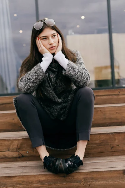 Stylish dark-haired city girl in a gray sweater sits on the steps, glasses on her head, thinks about something — Stock Photo, Image