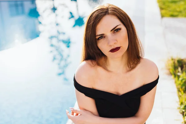 Una mujer joven sexy de lujo en un vestido negro se sienta junto a la piscina azul, disfruta de relajarse — Foto de Stock