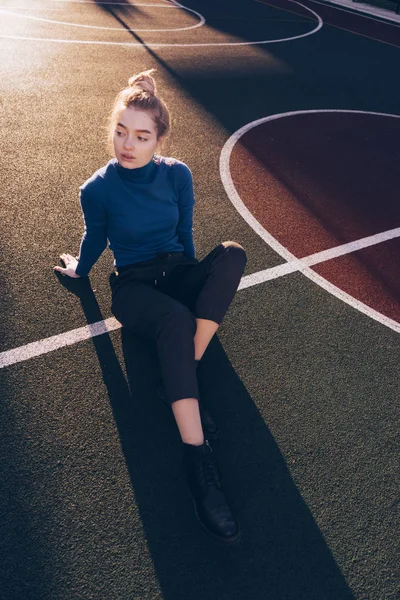 Delgada joven modelo en un suéter azul se sienta en un campo de deportes al aire libre, en los rayos del sol de la mañana — Foto de Stock