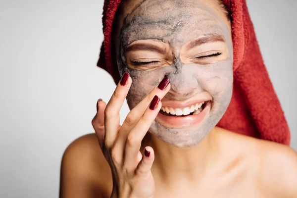 Счастливая смеющаяся молодая девушка с полотенцем на голове надевает полезную глиняную маску на лицо — стоковое фото