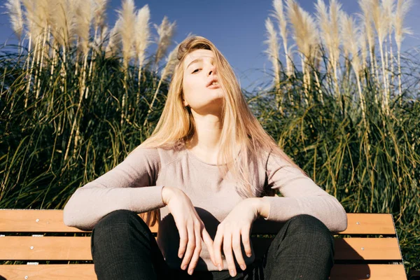 Cool ragazza sicura di sé modello biondo seduto su una panchina all'aperto, in posa al sole — Foto Stock