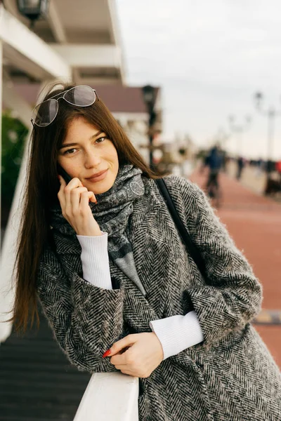 Красива темноволоса міська дівчина в сірому пальто, що йде вулицями міста, розмовляє по телефону — стокове фото