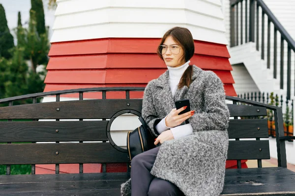 Élégante fille de la ville aux cheveux foncés en manteau gris et lunettes se trouve sur un banc à l'extérieur, temps nuageux — Photo