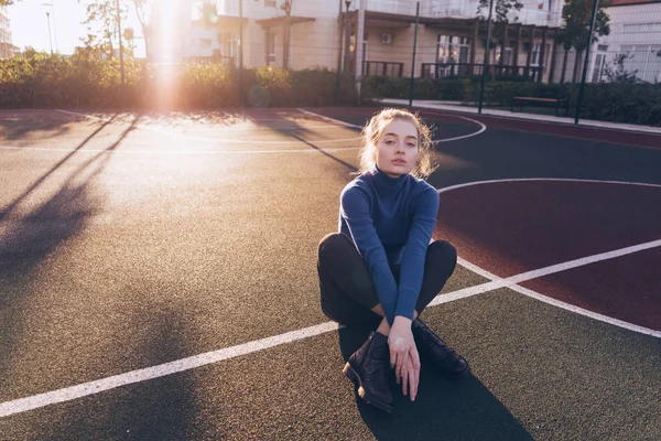 Elegante modelo de chica hermosa en un suéter azul se sienta en un campo de deportes en el sol — Foto de Stock