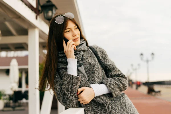 Стильна молода темноволоса міська дівчина в сірому пальто і окулярах розмовляє по телефону, гуляючи по вулицях міста — стокове фото