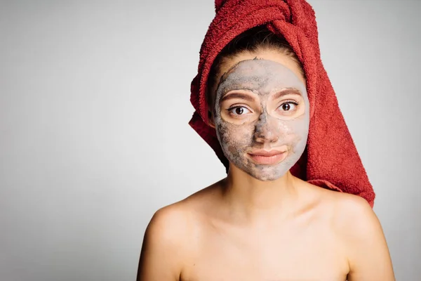 Vacker ung flicka med en röd handduk på huvudet tillämpas en helande lermask i ansiktet — Stockfoto