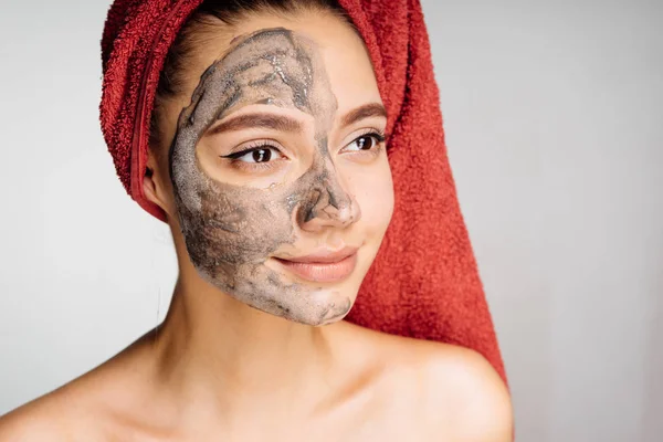 Молодая девушка с полотенцем на голове хочет быть красивой, надеть глиняную маску на половину лица — стоковое фото