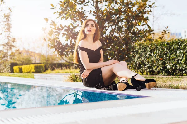 Una donna ricca di lusso attraente in un abito nero riposa vicino alla piscina blu, guarda pensieroso fuori da qualche parte — Foto Stock