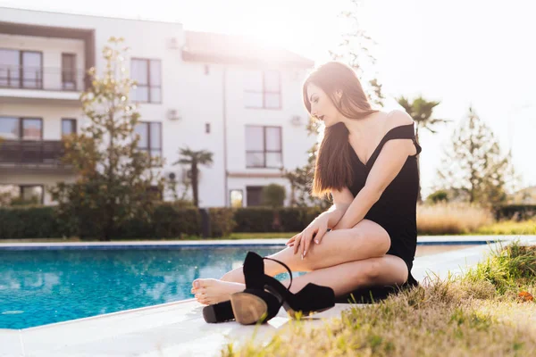 Σέξι κορίτσι ελκυστικό σε ένα μαύρο φόρεμα κάθεται δίπλα στην πισίνα, έβγαλε τα παπούτσια της, παίρνει ζεστό καιρό και τον ήλιο — Φωτογραφία Αρχείου