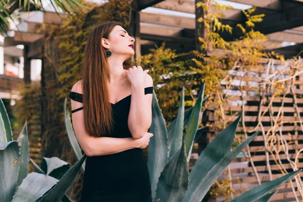 Sexy luxurious young woman in a black dress walking through her garden, enjoying nature — Stock Photo, Image