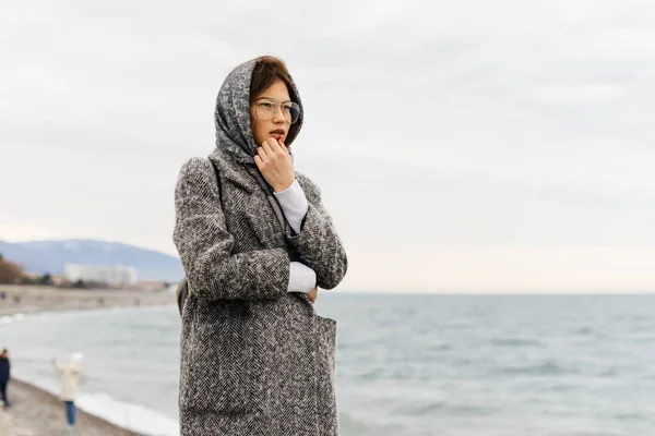 Dark-haired beautiful young girl in a gray coat and glasses walks by the sea, cloudy weather and wind — Stock Photo, Image