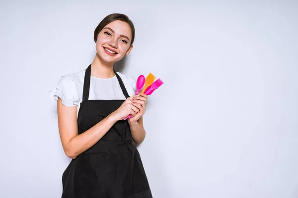 Усміхнена молода дівчина шеф-кухаря в чорному фартусі тримає інструменти для приготування і випічки — стокове фото
