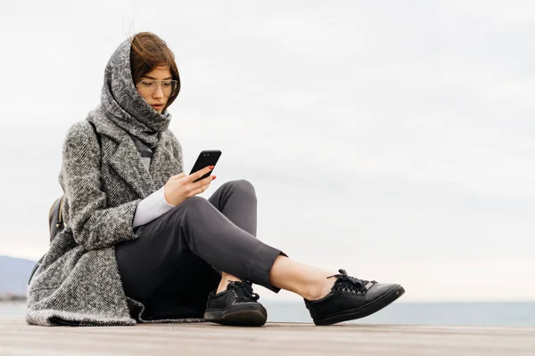 A stylish dark-haired girl in a gray coat and glasses sits by the sea, writes a message on the phone — Stock Photo, Image