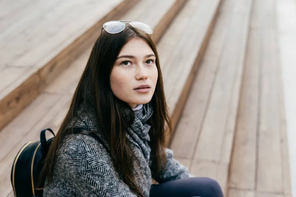 Beautiful young urban brunette girl sitting on the steps outdoors, looking up, street style — Stock Photo, Image