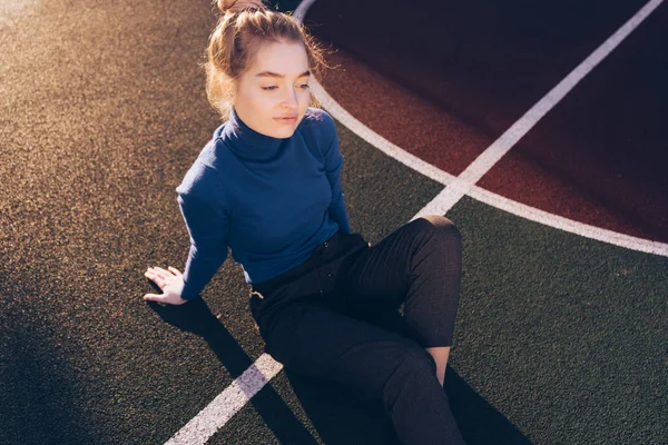 Stijlvolle mooi jong meisje in een blauwe trui zit op een sportveld poseren in de zon — Stockfoto