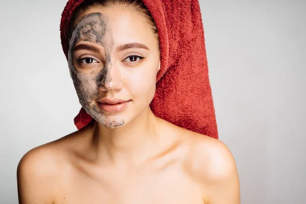 En ung vacker flicka med en röd handduk på huvudet tillämpas en användbar lermask på halva ansiktet — Stockfoto