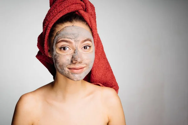 Ένα χαριτωμένο νεαρή κοπέλα με μια κόκκινη πετσέτα στο κεφάλι της εφαρμόζεται μια θρεπτική μάσκα αργίλου στο πρόσωπό της, την ημέρα του σπα — Φωτογραφία Αρχείου