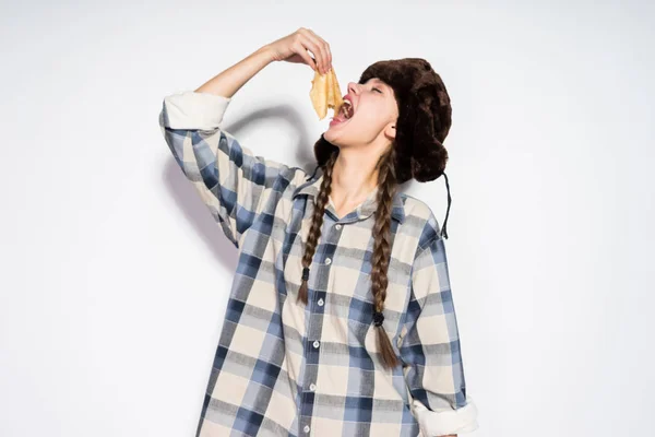 Una giovane ragazza russa in un cappello di pelliccia mangia frittelle calde deliziose, celebra Shrovetide — Foto Stock