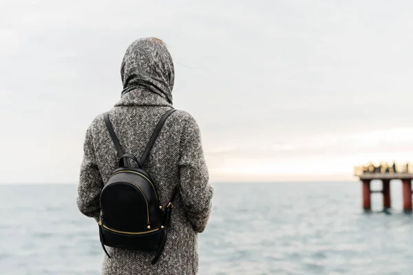 Stylish girl in a gray coat and with a black backpack travels, looks at the calm sea — Stock Photo, Image