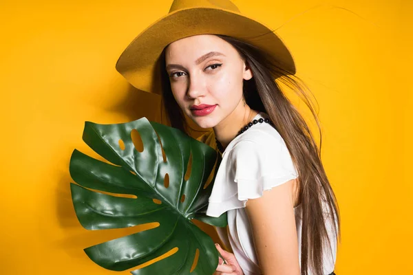 Confident stylish girl model in a fashionable hat holds a green leaf, posing on an orange background — Stock Photo, Image