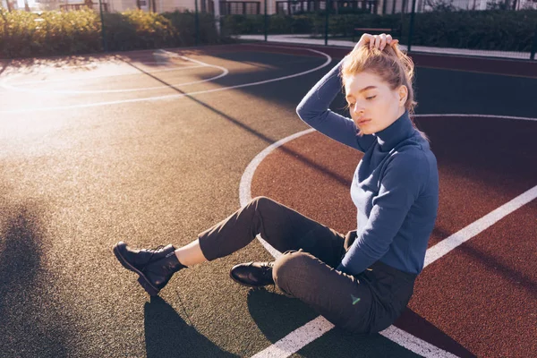 Stijlvolle jong blond meisje in blauwe trui zit op speelplaats in de ochtend zonnestralen — Stockfoto