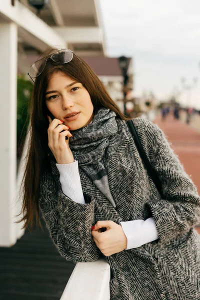 Bella ragazza di città dai capelli scuri che parla al telefono, passeggiando per la città — Foto Stock