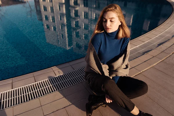Stylish young blond model girl in blue sweater sits by pool, enjoys the sun — Stock Photo, Image